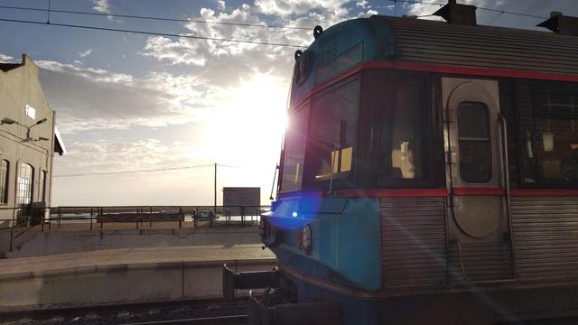Der Triebkopf eines portugiesischen Suburbano-Zuges an einem unübersdachtem Bahnhof. Das Foto ist im Gegenlicht der Sonne aufgenommen. Der Zugkopf füllt fast die komplette rechte Bildhälfte aus. Links sieht man ein altes, weißes Bahnhofsgebäude.