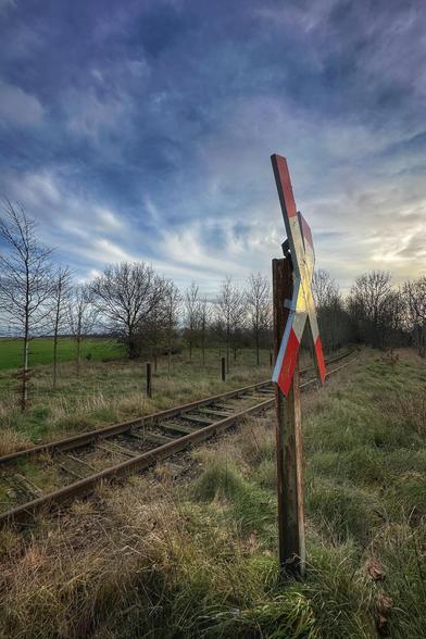 Von Gras überwucherte und rostige Bahnschienen verlaufen sich in der Ferne. Im Vordergrund ein Andreaskreuz, das von der Abendsonne angestrahlt wird. 