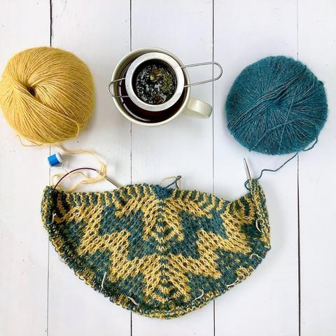 Flat lay view of a triangular shawl with mosaic patterning being knit on a circular needle. It’s shown with a ball of yellow and a ball of petrol colored yarn, and a cup of tea on a white plank background.