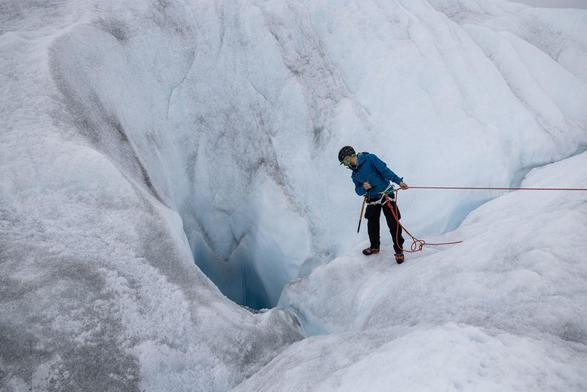 Person (with a safety rope) leaning over an icy landscape to look into a water-filled hole in a glacier