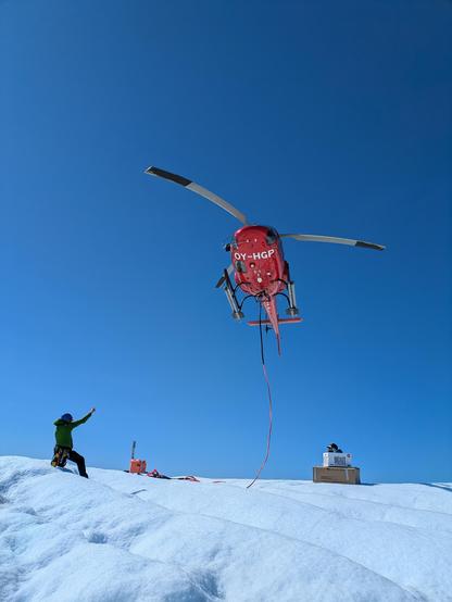 Red helicopter coming in to land against a blue sky. A person on the ice below is signalling to the pilot with a raised hand.
