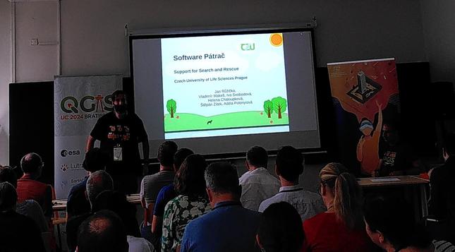 Low-quality foto (rainy wearher, low amount of light, etc.) of Search and Rescue Misson Support presentation by Jan Růžička.

Jan along with his first slide, some posters along with backs of some attendees visible.