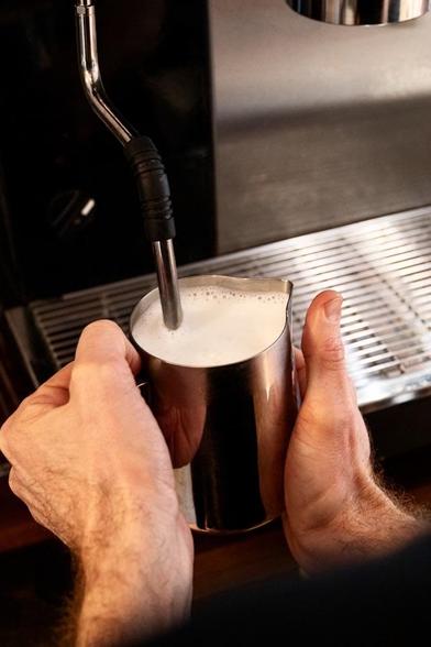 A close-up of hands holding a metal milk frothing pitcher with foamed milk inside, near a steaming wand from an espresso machine.
