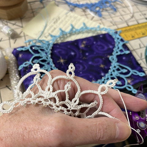 Tatting partially finished, in white thread held in a hand, with unfinished tatting in blue thread in the background.