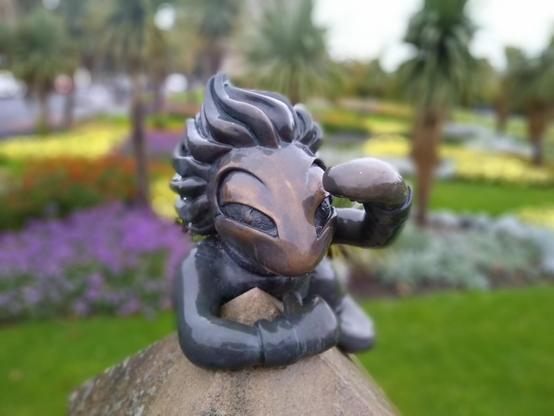 A bronze cast of a lemming from the computer game "Lemmings" looking directly at the viewer, left hand shading their eyes, with their right hand clinging to a pyramid-shaped stone top. In the background blurred trees that look vaguely like palms and flower beds.