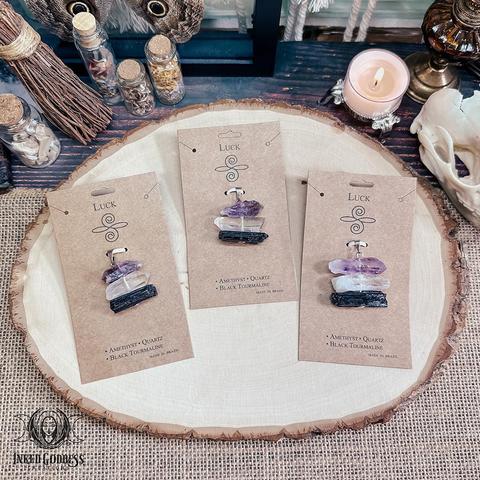 Three specimens of the "Luck Raw Gemstone Pendant" product are arranged on a raw wood slice. They consist of three small rectangular specimens of amethyst, clear quartz, and black tourmaline strung atop one another and attached to a silver pendant bail. They are packaged on brown cards labeled "Luck" and the names of the three gemstones. Presented by Inked Goddess Creations.