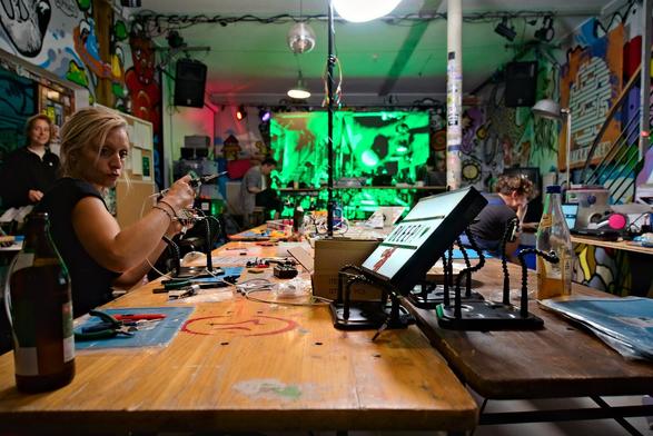 interior shot of the venue. people sitting at a table soldering equipment. in the background a screen with video art