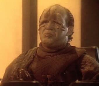 Alien guy from TNG with some face loaf who looks dumber than a box of hair is standing and looking blankly.  He's wearing a tattered potato sack looking frock. No caption. 