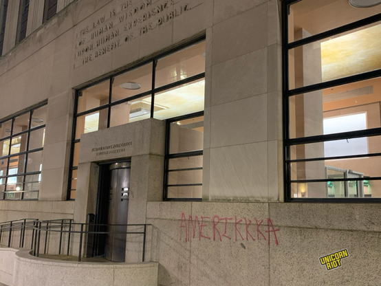 “Amerikkka” written in red all capital letters in spray paint on the stone wall just to the right of the rounded off entryway in front of thick turnstile style brown metal doors with thin window slots in them by large glass windows in the front of the US Post Office and Passport Office in center city Philadelphia at night