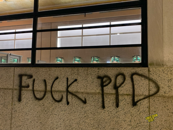 “Fuck PPD” written in all caps in black spray paint on a stone wall just below the US Post Office/Passport Office window through which a well-“85 lobby is visible with about a half dozen green triangles seen protruding from the walls which read “All Services”