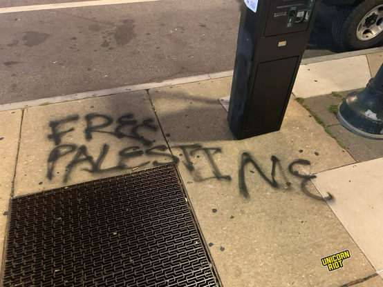 “Free Palestine” written in all caps in black on a sidewalk close to the street in between a square venting grate on the bottom left and a computerized parking meter and street lamp base on top right