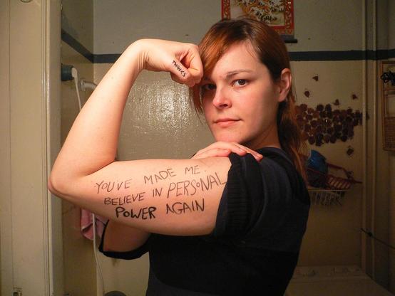 Photograph of a woman in a black tee shirt from the side. Her left upper arm is parallel to the ground, her forearm is bent at the elbow and her hand is bent back touching her head with her fist clenched. On her thumb is written the word 