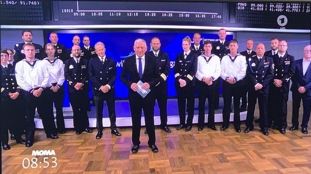 Eine Gruppe von Menschen, darunter Militärangehörige in Uniformen und ein Mann im Anzug, ist in einem Studio versammelt. Der Hintergrund zeigt einen blauen Bildschirm mit Text. Die unten angezeigte Zeit ist 08:53.
Börsenparkett Frankfurt.