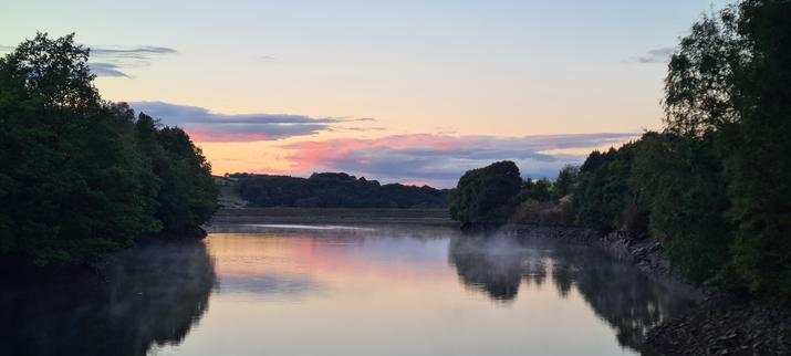 A narrow reservoir, with mature broadleaf trees on each bank. In the distance, there are clouds glowing in pinks and golds courtesy of the sunset - and the colours are reflected in the still water below.