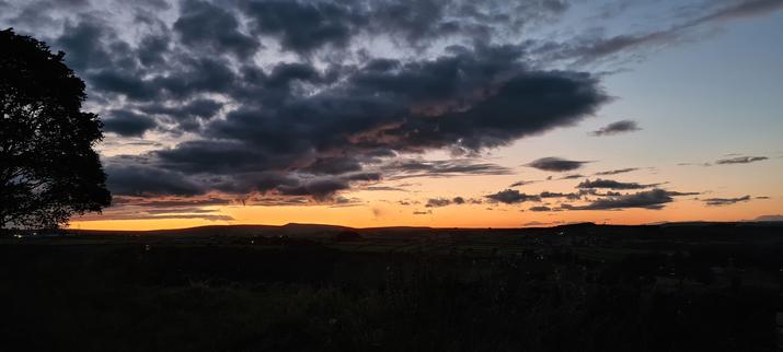 A wide, hilly landscape at sunset. The land is in shadow as are some menacing clouds above, but there is a golden orange glow on the horizon behind the hills.