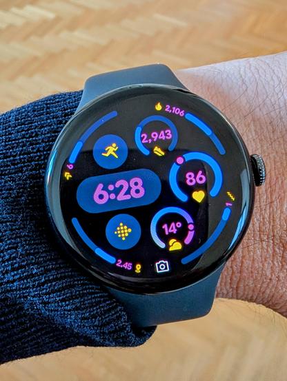 A close-up photo of a Google Pixel Watch 3 on a person's wrist. The watch face displays the time as 6:28, along with various fitness and weather information, including step count, heart rate, and temperature.