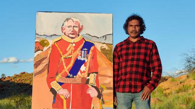 Vincent Namatjira's October 2024 show, Desert Songs, will be displayed at the Yavuz Gallery in Sydney.
Source: AAP / Jesse Lizotte

Image Portrait painting of King Charles in royal attire with medals monarch sash etc out in desert country with mountain range behind. 
Next to painting is The artist VN wearing blue jeans and a red and open neck black and red plaid shirt.
Behind painting and its artist is red dirt/yellow rock country and blue sky.