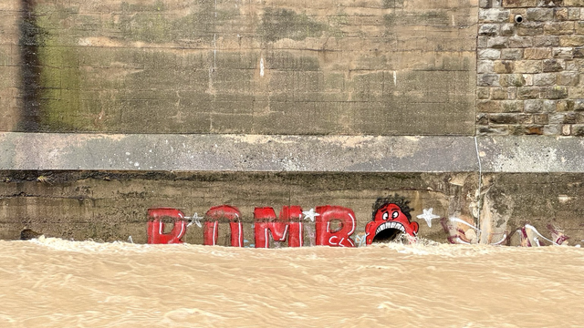 Floodwater flows through the city in an old brick trench. A pipe with a diameter of 40 centimeters emerges from the wall. Someone has painted a fiery red head around it, turning the pipe into a wide open mouth. A row of teeth can be seen at the top, but the lower half is completely surrounded by the extremely high water level. Normally it looks as if water is flowing out of the mouth, but now it looks as if this figure is swallowing the water and threatening to drown.