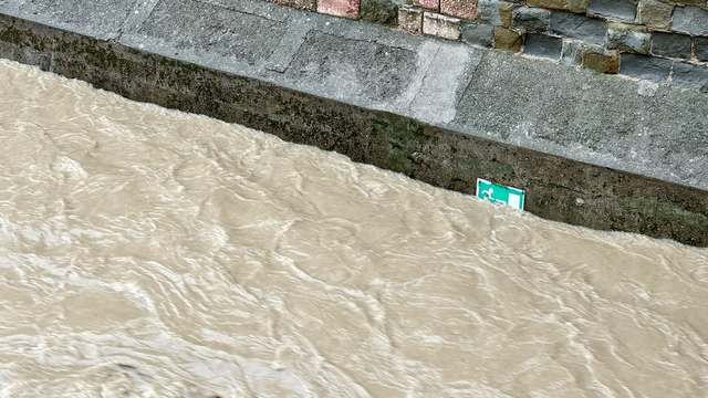 Floodwater flows through the city in an old brick trench. A white and green sign for an emergency exit is already half submerged.