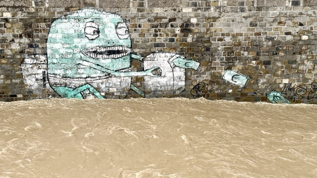 Floodwater flows through the city in an old brick trench. A graffiti shows an egg-shaped green and white creature in white underpants. It sits there with its skinny legs bent. With its scrawny little arms, it operates a button on a box-shaped device, probably something like a money printing machine. Several green banknotes fly in a wide arc into the rushing floodwater.
