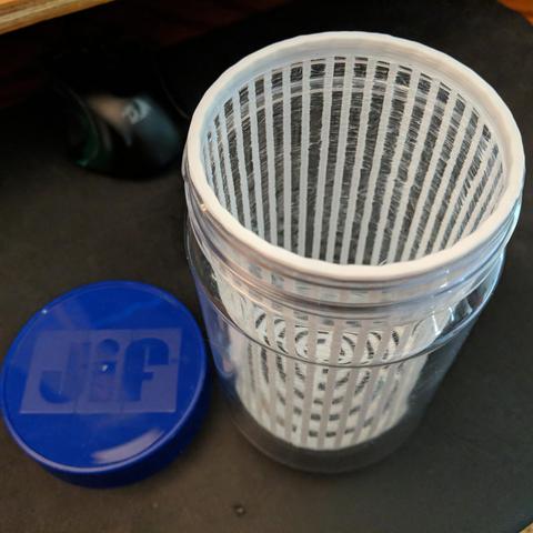 The prototype strainer in the plastic jar. It was a single wall thickness and very stringy and slightly oversized spacing because the thin columns moved as they were printed. The blue lid for the jar sits to the left and it says jif on the top. If it leaks a lot two TPU gaskets can be printed. One under the strainer lip and one to fit in the lid. It is printed with PLA. The wash for the bees will use soap, not alcohol. 