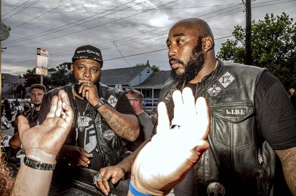 Bikers in black leather. Two men in the foreground, more in the background, and the shot includes another man’s outstretched hands.  It’s a moment in a conversation.