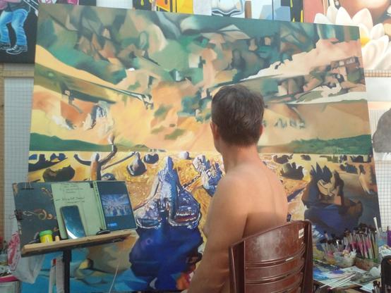 a photo of Kem, his back to me, looking at his easel, an ipad next to him with the piece of digital art on it, the painting is a large strange abstract scene, a big dreamlike landscape with lots of abstract shapes and figures 