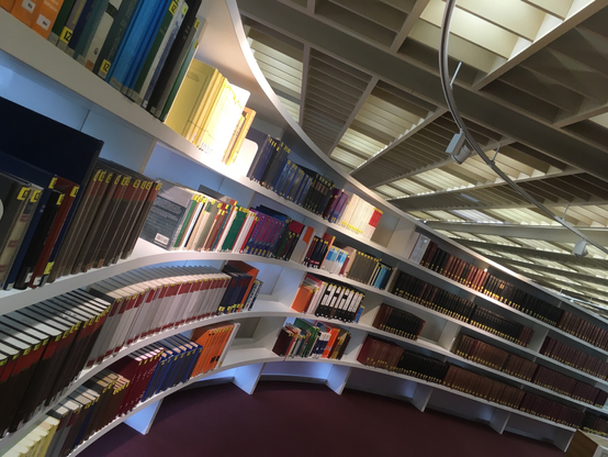 Ein weißes Bücherregal etwas schräg fotografiert. Bücher unterschiedlicher Farben und Größen stehen darin. Das Regal verläuft gebogen durch einen großen Raum.