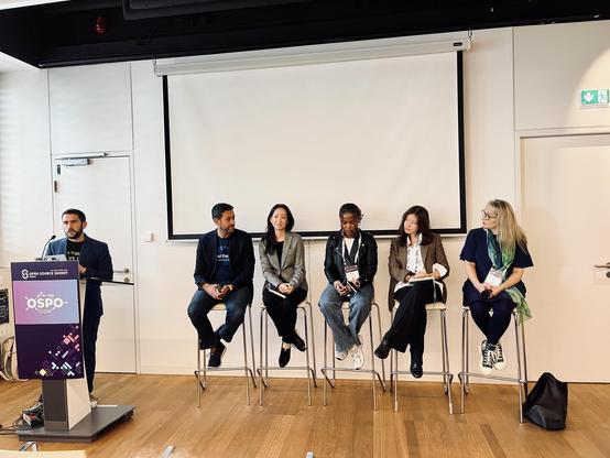 Panel at Open Source Summit Europe in Vienna. Nick Gates is standing behind a podium with a sign "OSPO" while Omar Mohsine, Sachiko Muto, Ruth Ikegah, Fiona Krakenbürger, and Hilary Carter, sit on tall chairs in front of a white screen.