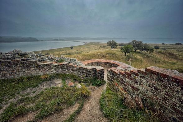 Erhöhter Blick vom verfallenen Gemäuer des Schlosses Kalø auf der gleichnamigen Insel  auf den Damm, den die Insel mit dem Festland verbindet. Auf der Insel stehen in der Ferne noch einige Bäume, die Sicht ist diesig, der Himmel dunkel, grau und verregnet. 