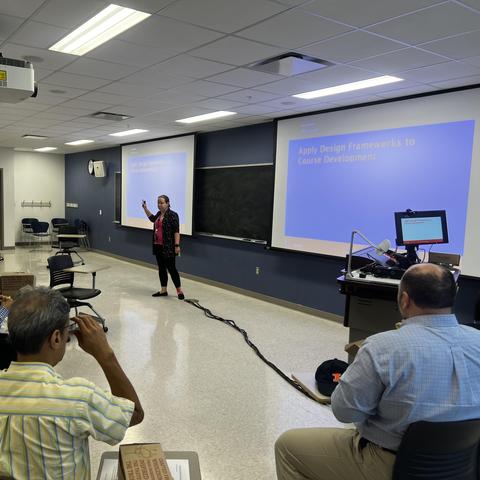 Two men are sitting at desks watching a woman give a presentation in a classroom with two screens showing the same slide. 