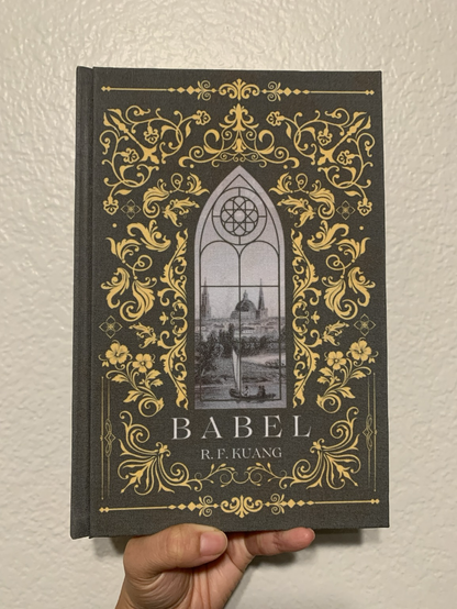 Image 1 shows a hand holding aloft a book titled Babel by RF Kuang. The bookcloth of the cover is green, with numerous filigree yellow designs surrounding a gothic arch window in the center. The window contains a steel plate engrave image of early 19th century Oxford, UK.