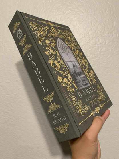 Image shows the same book now tilted at an angle to show the spine which reads "Babel" by RF Kuang, with more yellow filigree designs. Towns End Bindery's logo of a house on fire is shown at the top of the book spine.