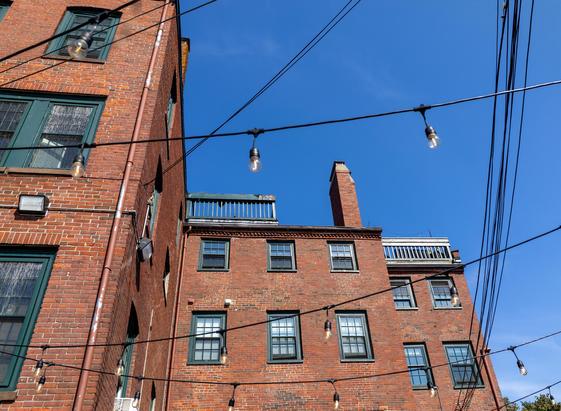 Color photo of part of a brick building and the blue sky above it. The angle of view and the roofline of the flat-roofed building form a zig-zag separation from the sky. The building has many windows with green painted frames and mullions. There are Edison lights strung across the image between the building and the camera position. There are also various utility wires haphazardly strung across the image.