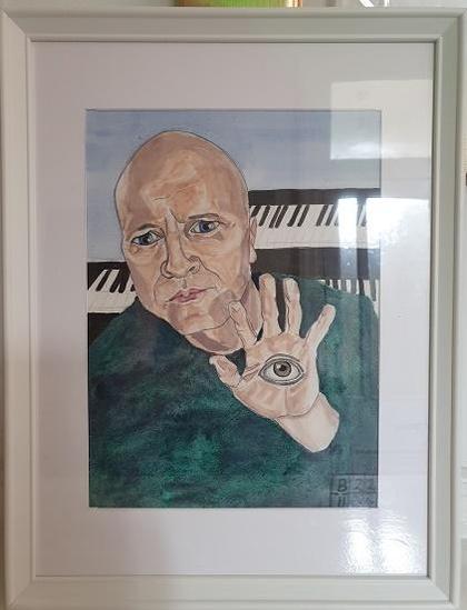 Id 1st image - portrait of Ivan in front of keyboards holding his hand up which has an eye painted on it. He is wearing a grey/green jumper and is bald.