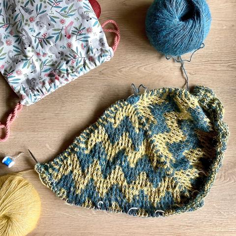 Flat lay view of a triangular shawl with mosaic patterning being knit on a circular needle. It’s shown with a project bag, and a ball of yellow and a ball of petrol colored yarn on a light wooden background.