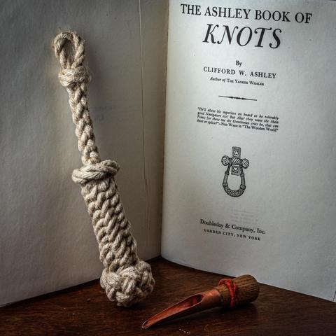 A photograph of a bellrope leaning against the Ashley Book of Knots, next to a copper and oak Swedish-style fid. 
