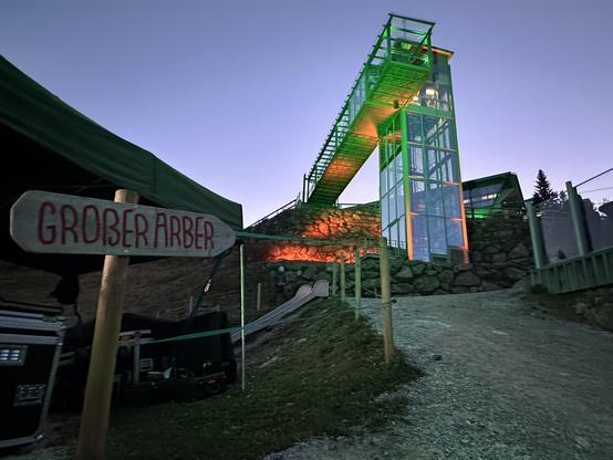 A glass and steel elevator with green lighting in front of a rocky cliff illuminated in orange. A gravel track leads towards it and there's a sign "Großer Arber" in the foreground.