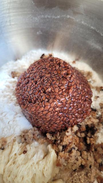 Farbfoto: Blick in eine Edelstahl Teig Schüssel, in der Mehl, eingeweichtes Altbrot, Sauerteig etc. zu sehen sind. Obendrauf liegt kopfüber das Leinsamen Brühstück, das gut erkennbar noch die Form des Gefäßes hat, in dem es gebrüht wurde. 