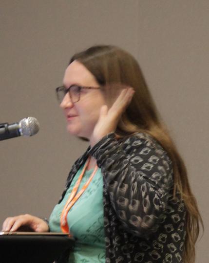 Kristina Collins, scientist of very long, very flowing hair, with her hand raised, communicating science, standing at a speaker's desk. She seems (and is) very enthusiastic about what she's talking about.