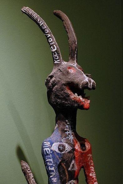 Figura humanoide con cabeza animal y largos cuernos curvos hacia atrás sobre su cabeza. Tiene pintado texto sobre los brazos, cola y cuernos y dos grandes ojos sobre el pecho.