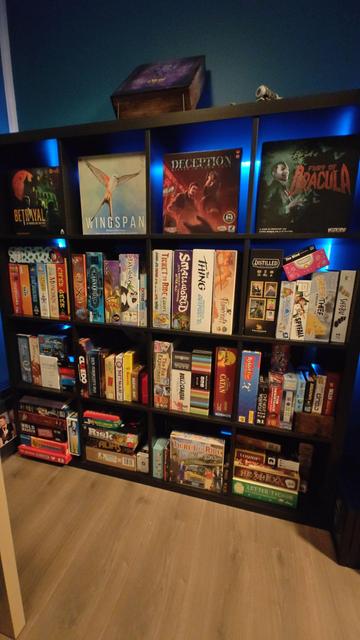 Bilete av ein reol fylt til randen av brettspel, med titlar som Blood on the Clocktower, Wingspan, Takenoko, Pitchstorm, Kast grisene, mfl. Reolen har eit blått lys som lyser opp veggen bak seg.