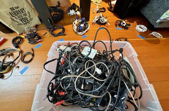 A chaotic collection of various cables and cords is shown, with a clear plastic container overflowing with tangled wires at the center. Surrounding it are neatly organized bundles of cables labeled on blue tape.