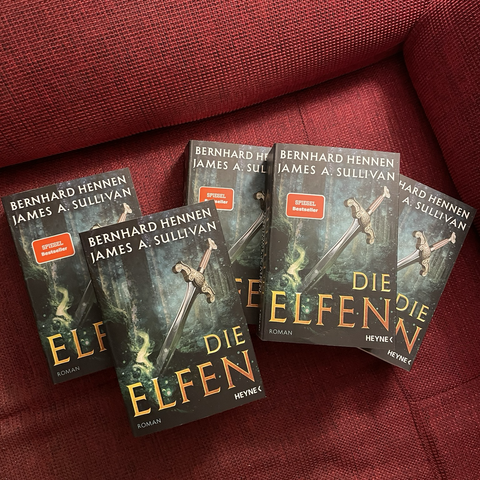 Fünf Exemplare der neuen Ausgabe von »Die Elfen«. Motiv: Schwert steckt in einem Waldboden. Titeln in goldener Schrift unten. Die Namen der Autoren – Bernhard Hennen, James A. Sullivan – oben. 