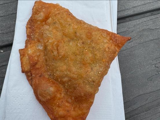 Photo of a samosa on a white paper napkin. the napkin is lying on a wooden picnic table. 

