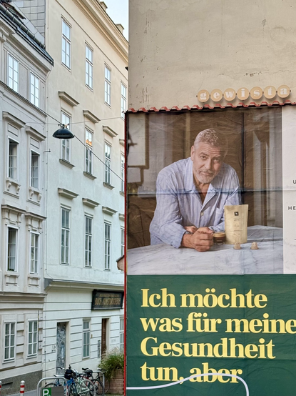 George Clooney in pyjamas, looking pensively from the front of a house in the old town, unable to make a proper coffee by hand. Underneath is the text from another advertisement: “I want to do something for my health, but”.