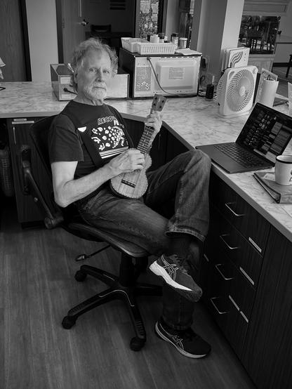 A photo of me behind aa coffee counter. Ukulele in my hand and a computer on the counter.  I am sad and the photo shows it