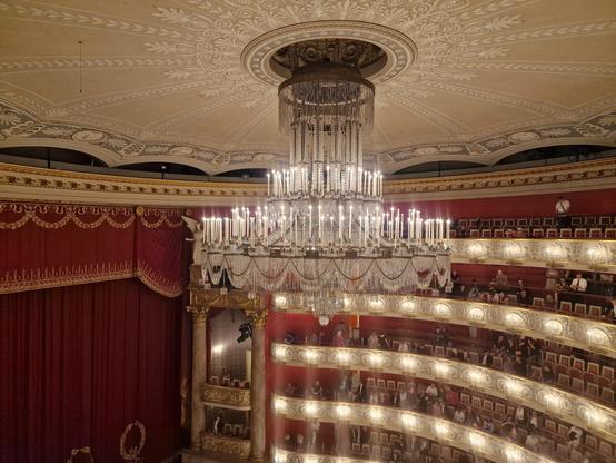 Chandelier in the opera, lit, with red, gold, and white decoration in the background.