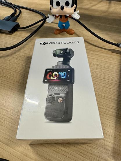 White box with “DJI Osmo Pocket 3” written at the top and showing a picture of the named camera, a skinny cuboid with a small lens unit on a gimbal at the top and a widescreen display on the body sticking out over the edges. “Osmo” is written just below the display and there is a joystick and a button with a red circle on it beneath that. A Goofy Funko Pop looks over the box