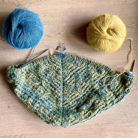 Flat lay view of the wrong side of a triangular shawl with mosaic patterning being knit on a circular needle. It’s shown with a ball of yellow and a ball of petrol colored yarn, on a light woodenbackground.